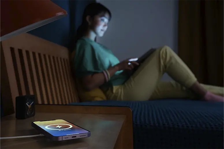 Un téléphone est posé sur une station de charge par induction pendant la nuit ; en arrière-plan une femme est assise sur un lit